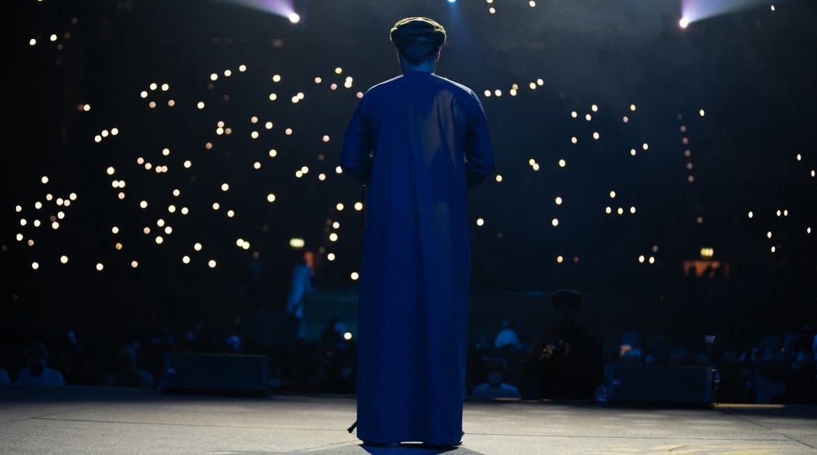 A speaker stands on stage facing an audience holding up phone lights in a darkened hall.
