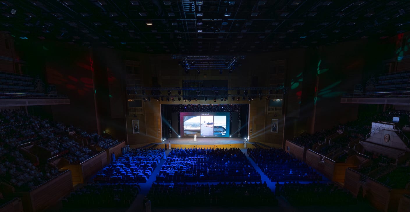 A full auditorium under blue light faces the stage at the closing evening.