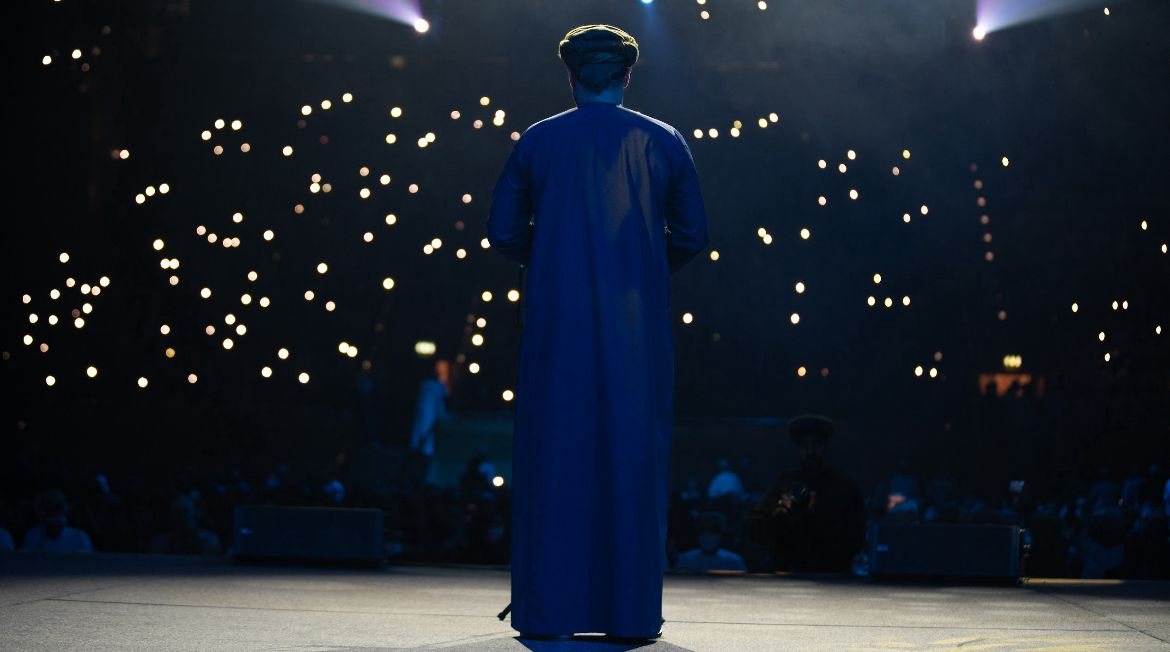 A speaker stands on stage facing an audience holding up phone lights in a darkened hall.