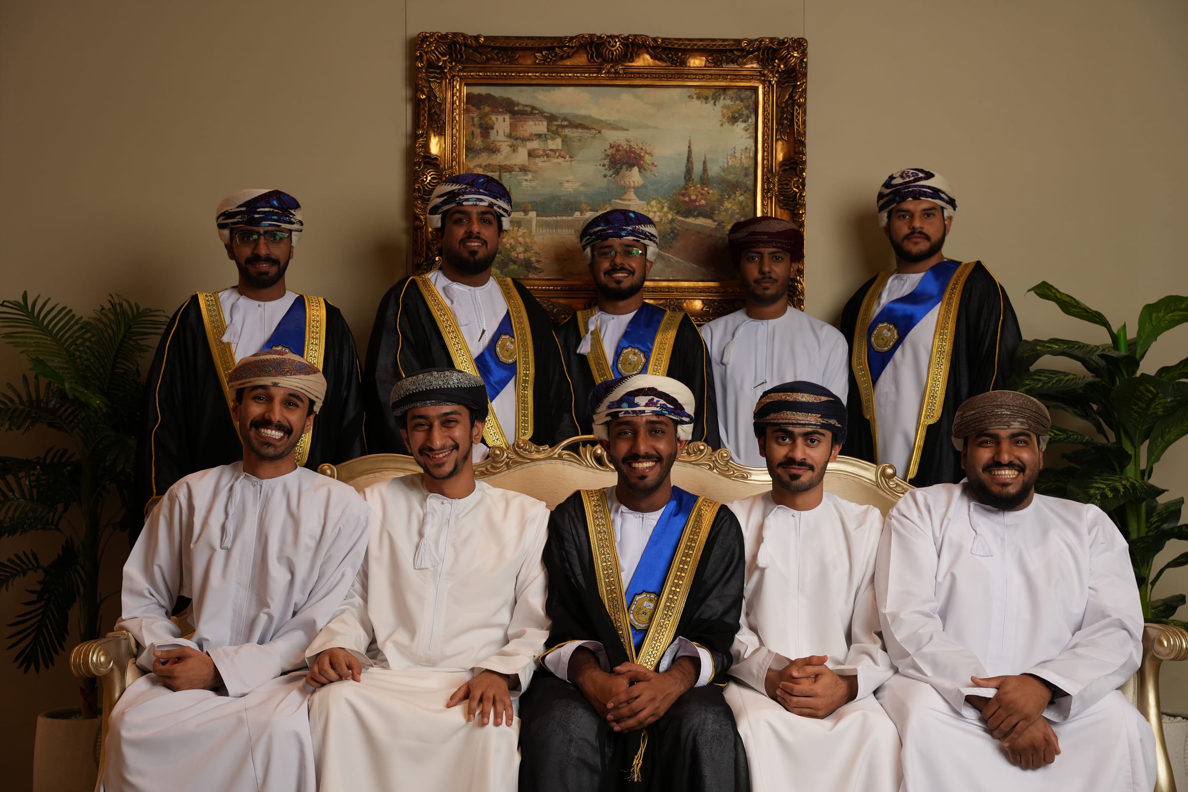Eleven graduates in white dishdashas and graduation bishts pose together for a studio portrait.