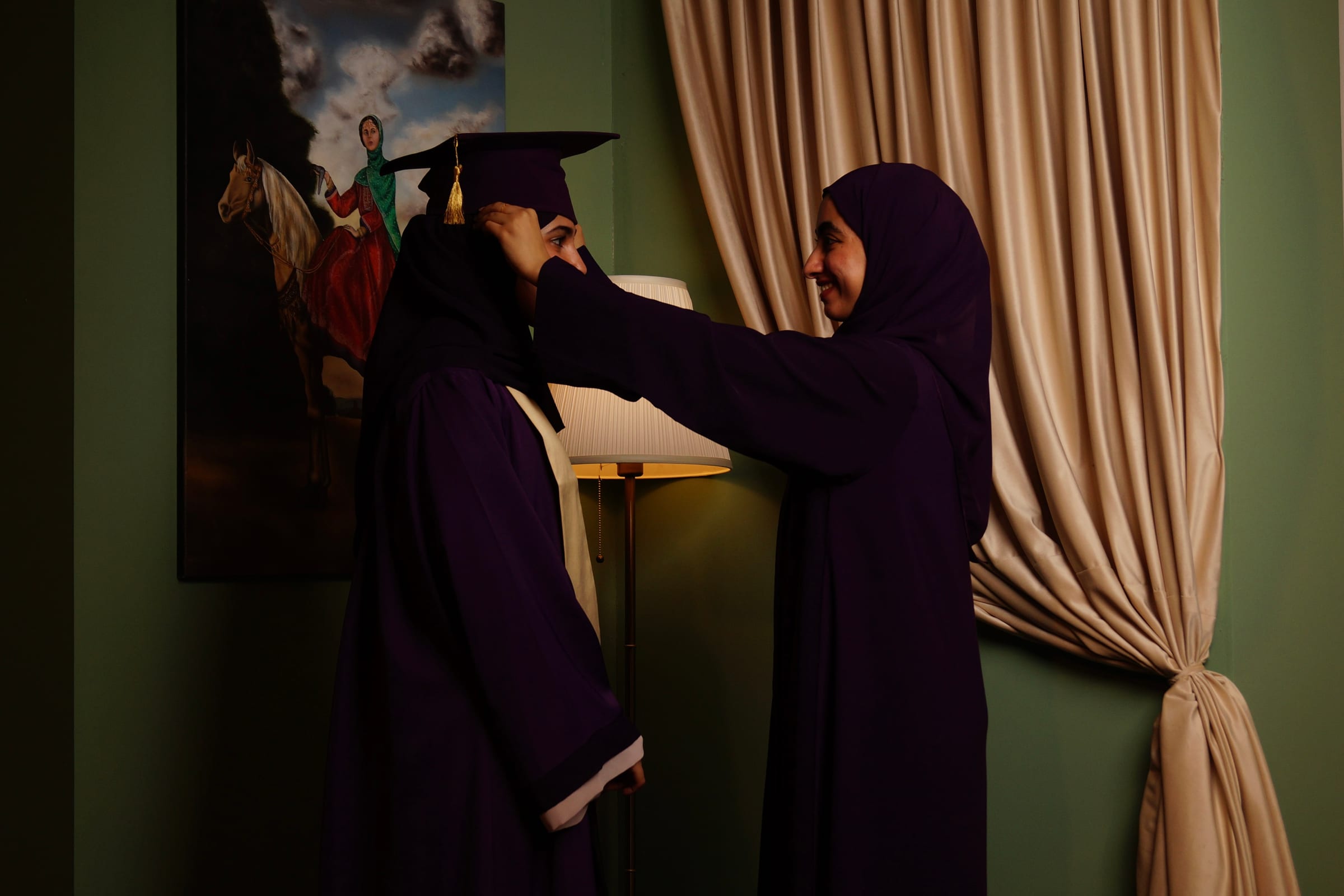 A friend adjusts the cap of a graduate in a purple gown, in a softly lit studio set.