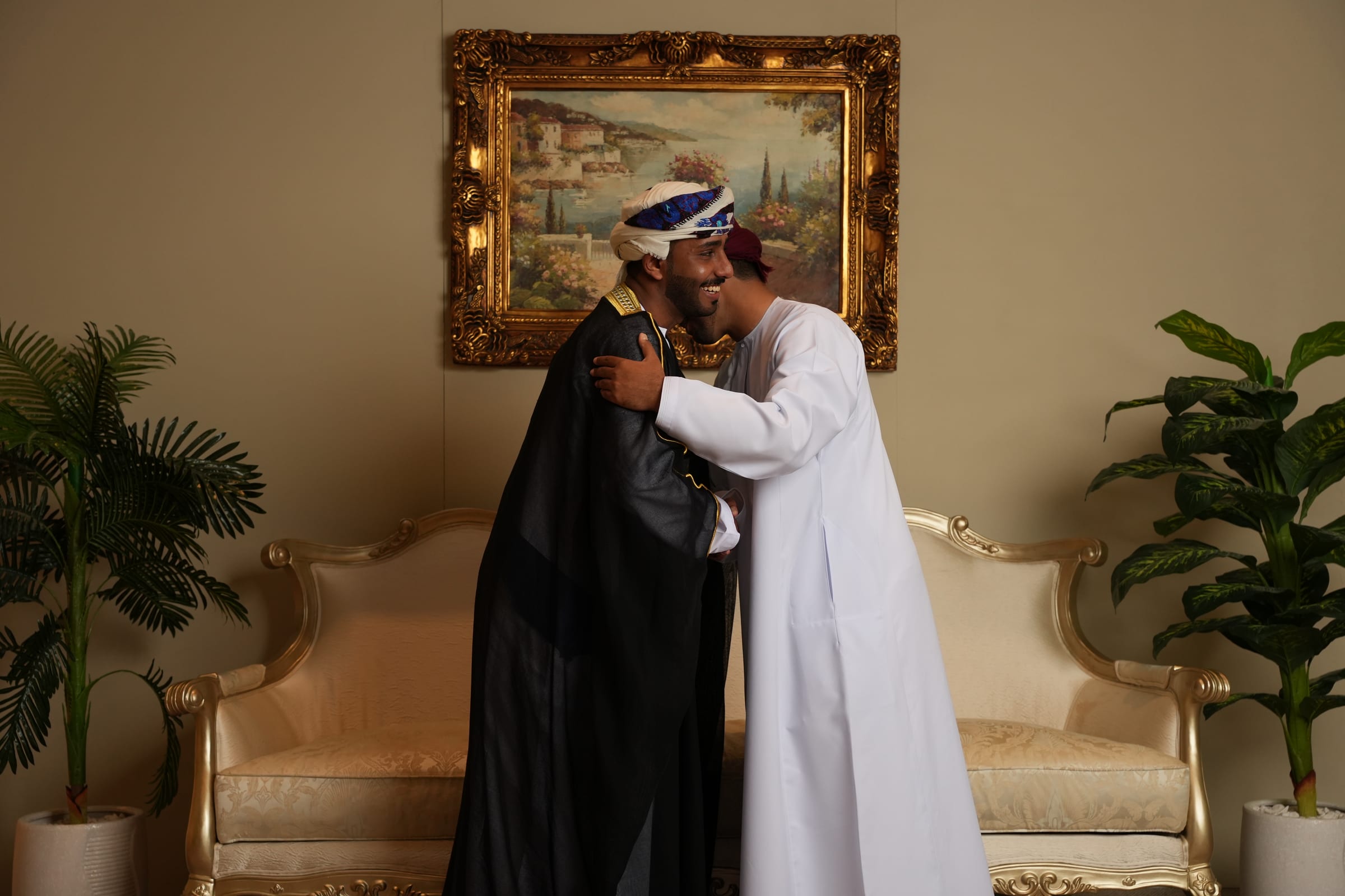 Two graduates embrace in front of a gilded sofa and a framed painting.