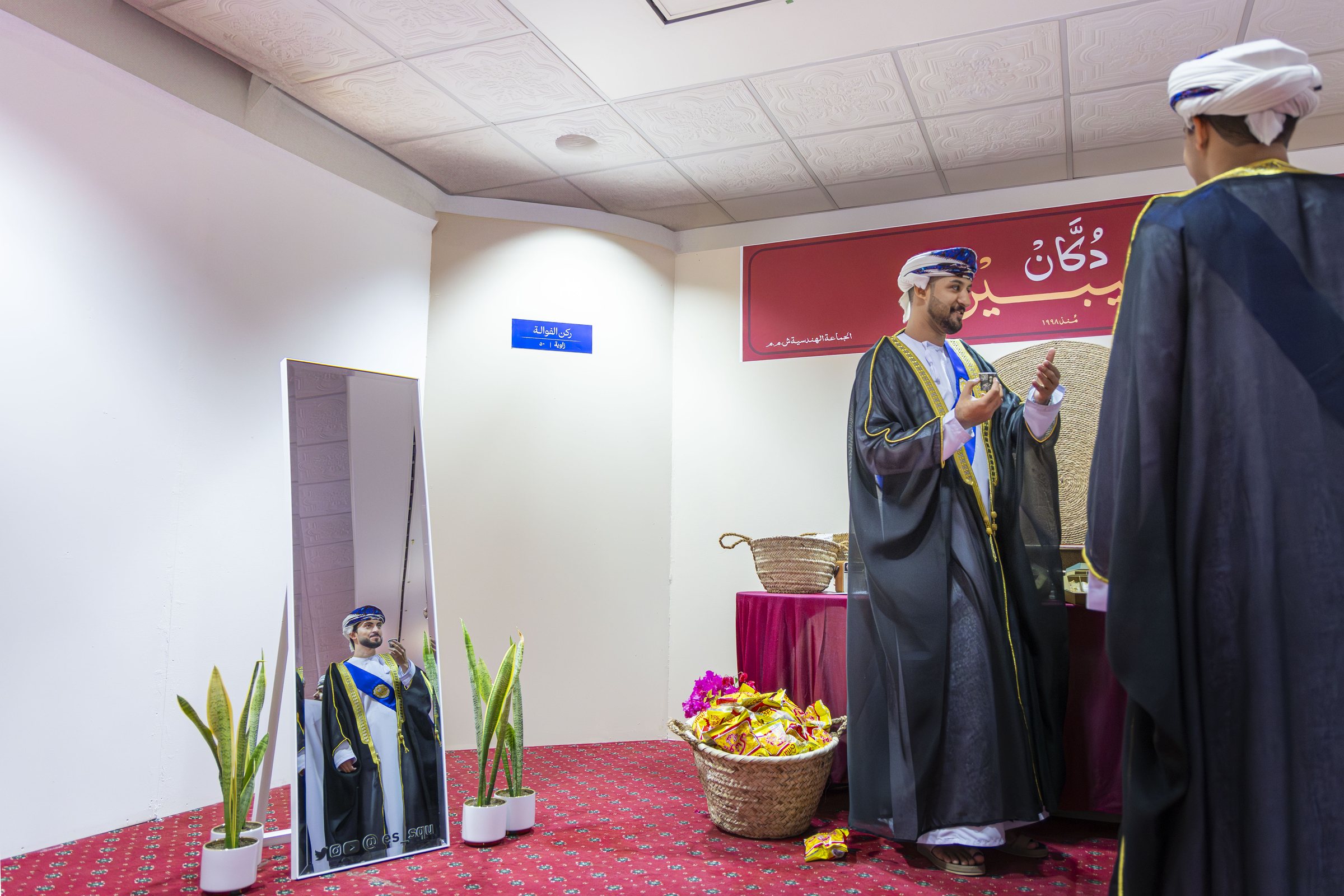 A graduate in a bisht poses in a decorated photo corner, his reflection caught in a standing mirror.