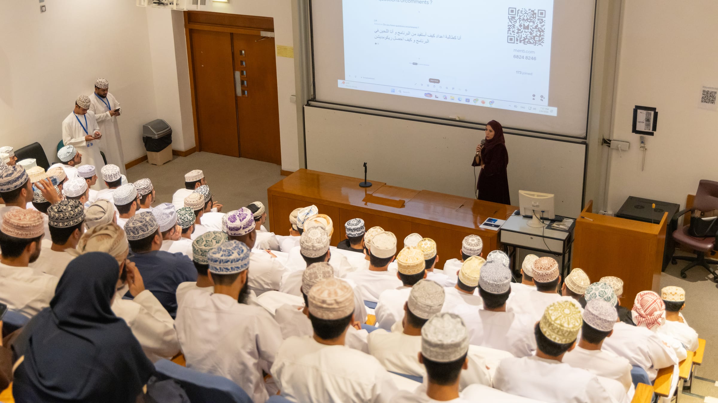 A speaker presents to a full lecture theatre of students, a question slide with a QR code behind her.